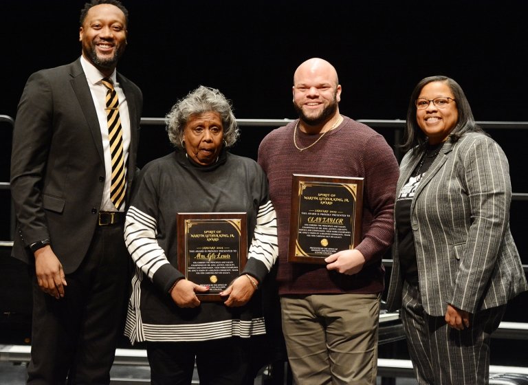 Two men and two women pose for a photo. A man and woman in the center of the photo hold awards they just received. 
