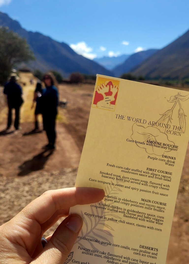 A hand holds a menu of Peruvian dishes with mountains and people seen in the background. 