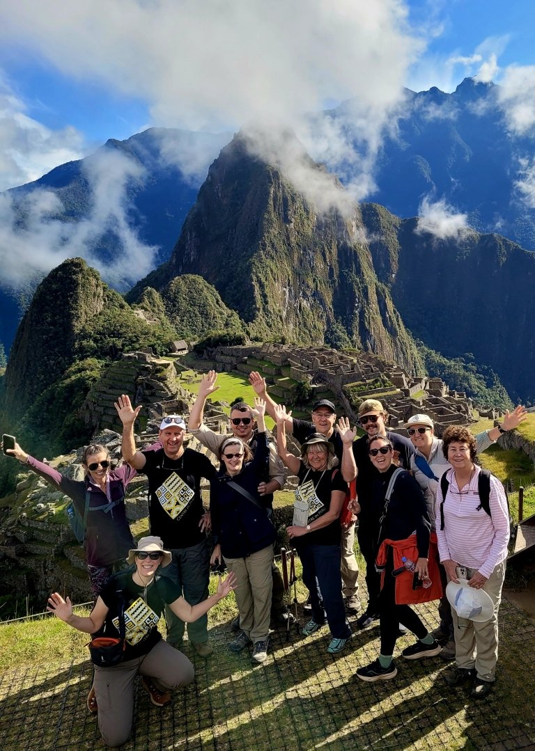 The steep mountains and stone dwellings of Machu Picchu can be seen in the background as a group of people pose for a photo in the foreground. 