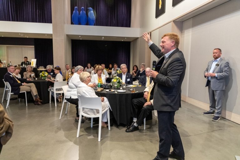 A man in a suit speaks into a microphone as guests seated at tables around a large banquet room look on. 