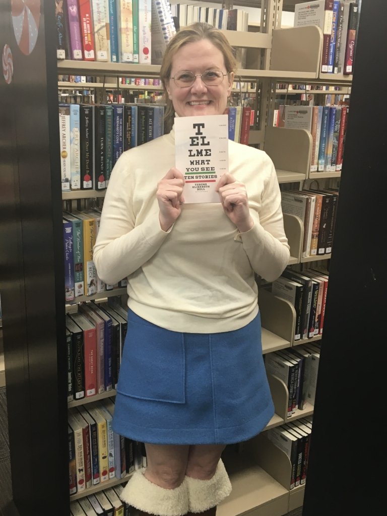 A woman holds a book in front of her while standing in a library between book-lined shelves. 