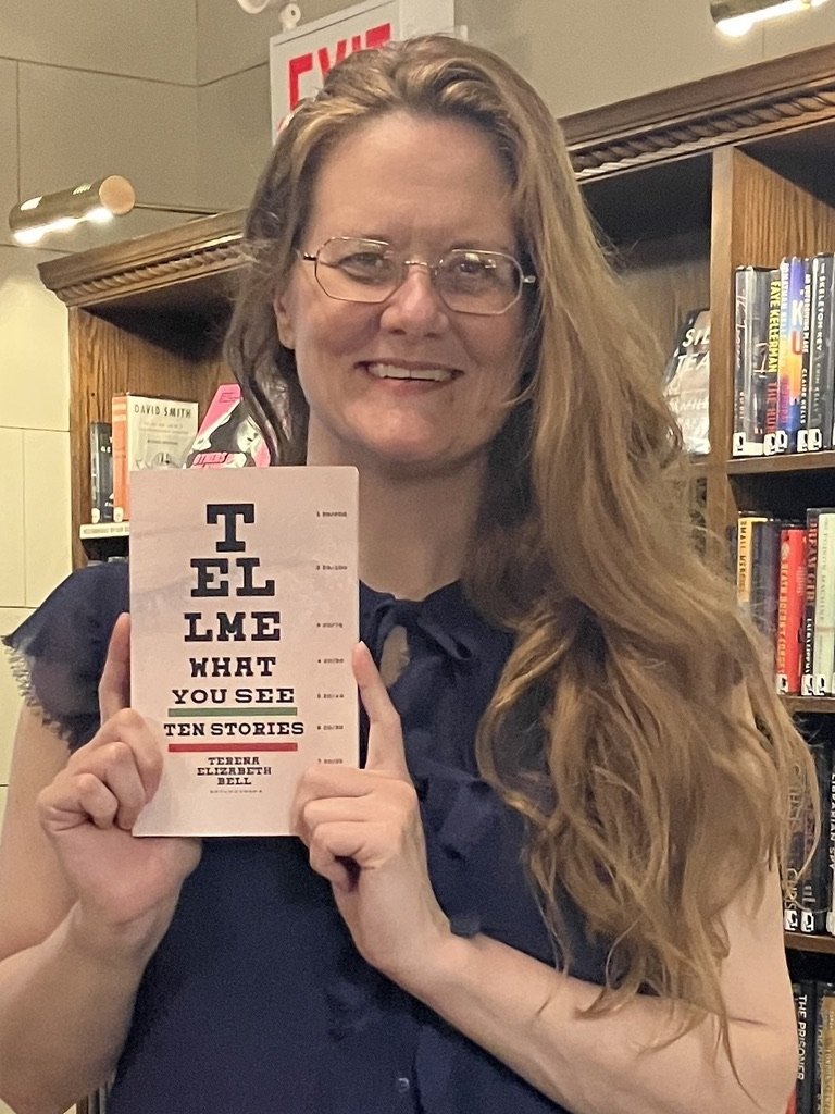 A woman with long hair wearing a dark shirt stands amid book-lined library shelves holding a copy of the book "Tell Me What You See."