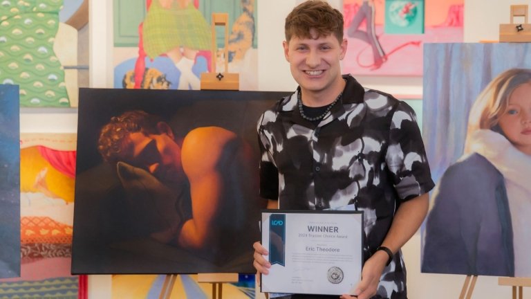 A man holding an award certificate stands in front of a painting of a person.
