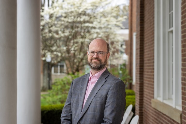 A man wearing a grey suit jacket and a pink shirt poses for a photo in front of a blooming dogwood tree. 