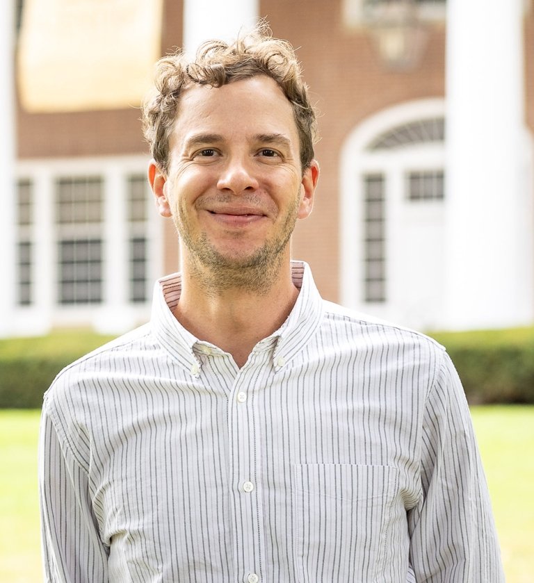 A man in a gray striped shirt poses for a photo in front of a brick building