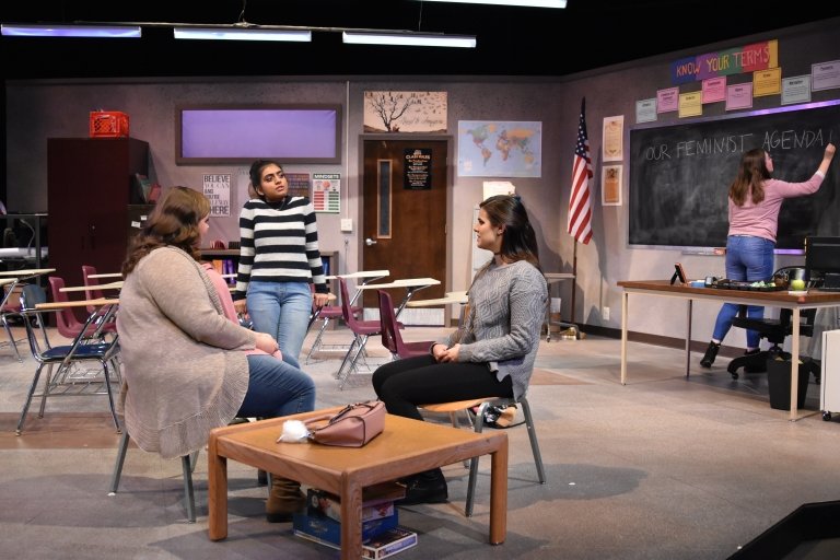 A stage set is designed to look like a high school classroom with three young woman talking while sitting in chairs in the foreground and another woman writes on a blackboard in the background.