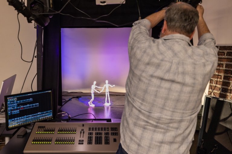 A man makes adjustments to a miniature theater light as two posable miniature figures are seen on a model stage.