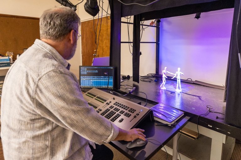 A man makes adjustments to a control panel as two posable miniature figures are seen on a model stage.