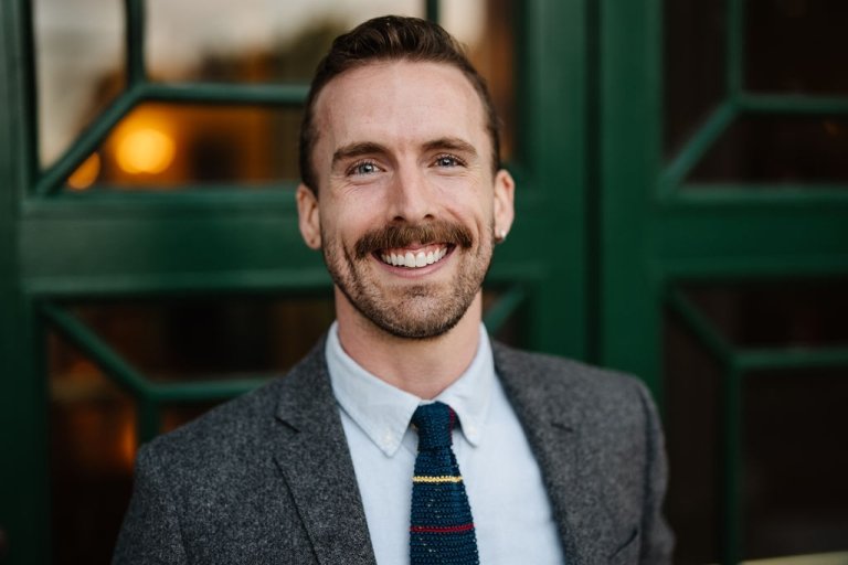 A man with a mustache and a dark suit and tie smiles as he poses for a photo. 