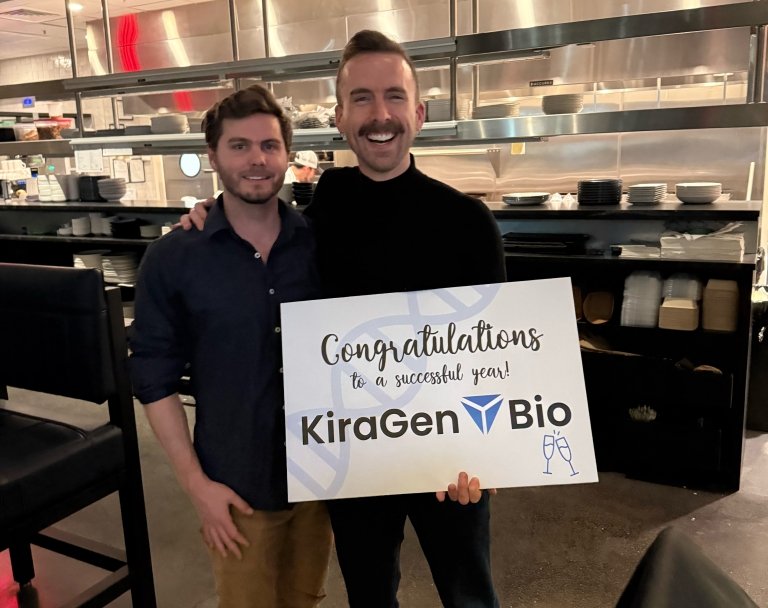 Two men post for a photo with a sign that states "Congratulations on a successful year KiraGen Bio."