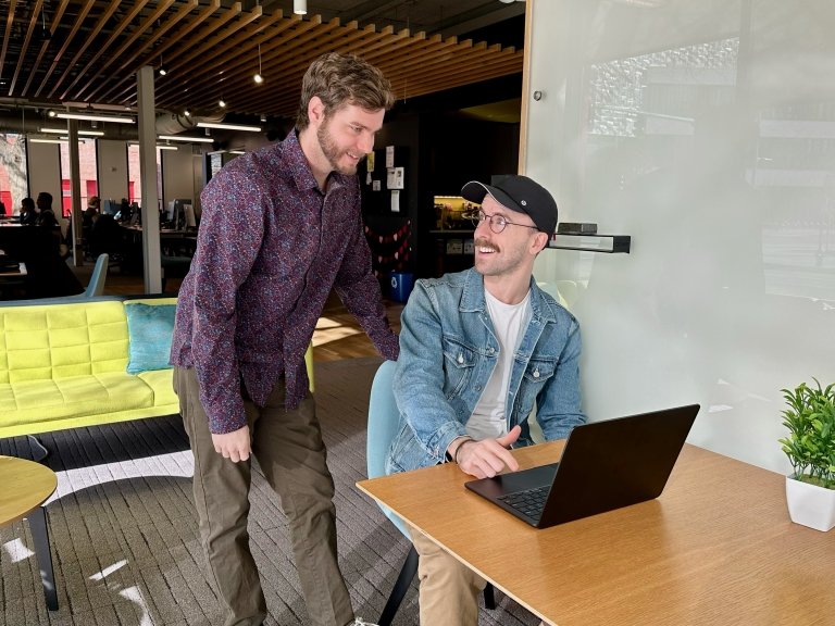Two men, one seated at a desk working on a computer and another standing behind him, chat. 