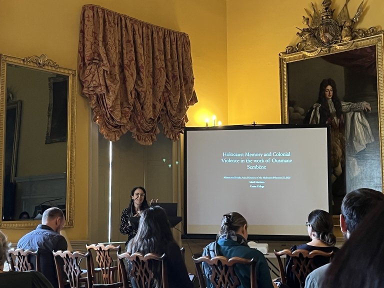 Six people sit at tables around a decorated room. They are looking at a projector screen with rows of small words on it.