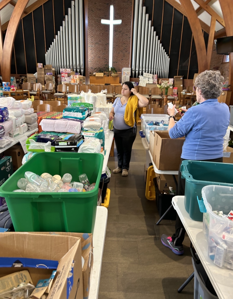A cross hands on the wall of a church sanctuary room which is filled with folding tables covered in boxes of donated goods. Two volunteers work with the materials.  