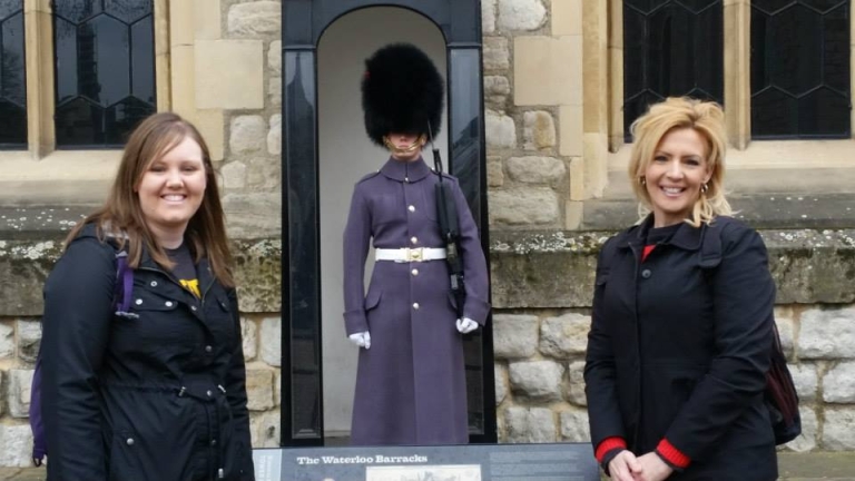 A young woman and her mother post for a photo with a royal palace guard visible in the background during a trip to London, England.