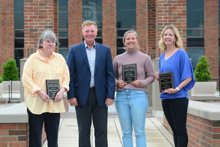 Three women pose with Centre President Milton C. Moreland for a group photo in front of a brick building while holding certificates of recognition.