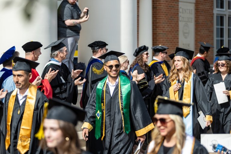 A student wearing a stole representing Brazil smiles in the procession line while being applauded by faculty and staff.