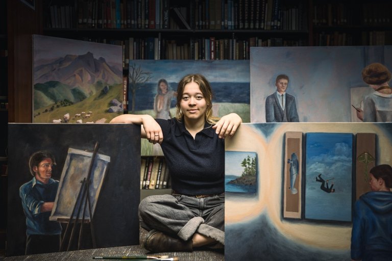 In a library setting, a young woman sits cross-legged on the floor surrounded by various paintings from her John C. Young project. The background is filled with bookshelves filled with books.