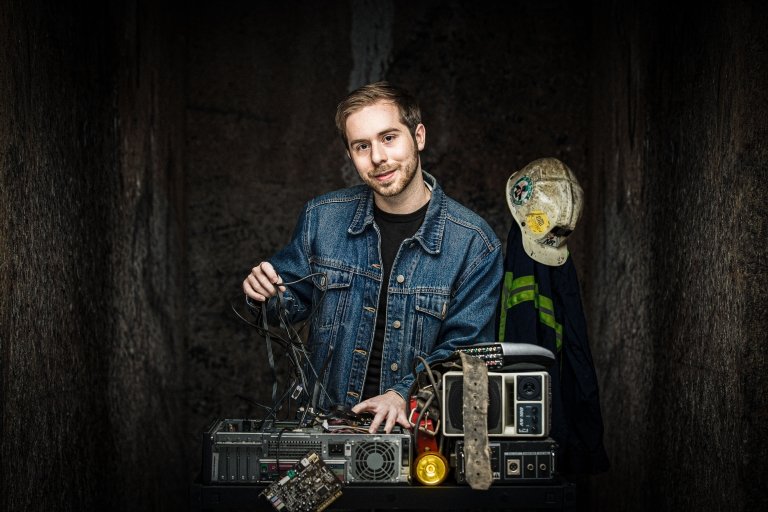 A young man stands in a dimly lit space, appearing to be in a cave. He is wearing a denim jacket over a black T-shirt. He has a confident smile as he holds several tangled cables in his right hand while resting his left hand on a vintage computer setup in front of him.
