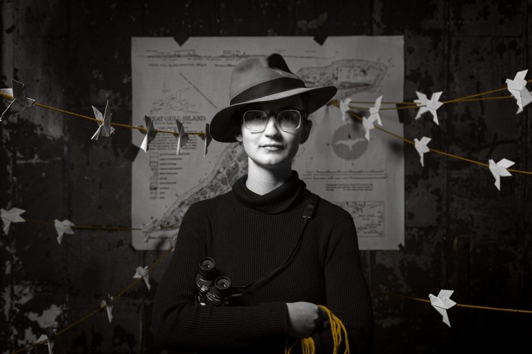 A person wearing a hat and glasses stands in front of a detailed map pinned to a dark wall. They hold binoculars in one hand and yellow string in the other, with paper cranes hanging from a line stretched across the background.