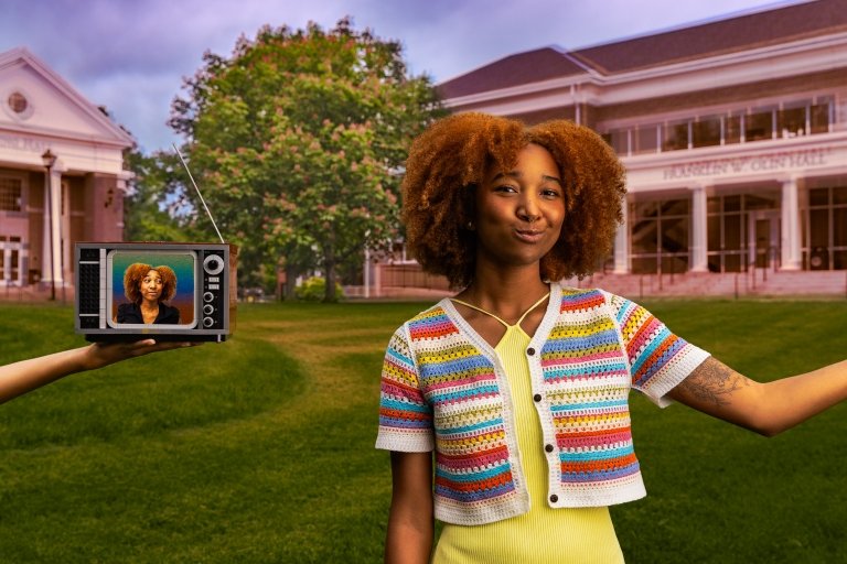 Kasey Montgomery, a young woman with curly, auburn hair, is standing on a green lawn in front of a large, classic academic building. In her left hand, she is holding an old-fashioned television set that displays her own image, mimicking her pose. The background includes a vibrant tree with pink flowers.