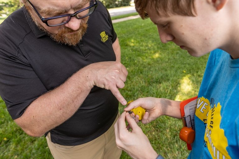 A student in a blue t-shirt holds a cicada shell as a man in a black polo shirt points to part of the shell.