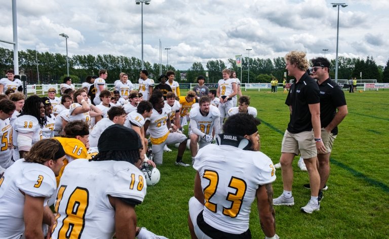 Two men in black speak with a group of football players on the field.