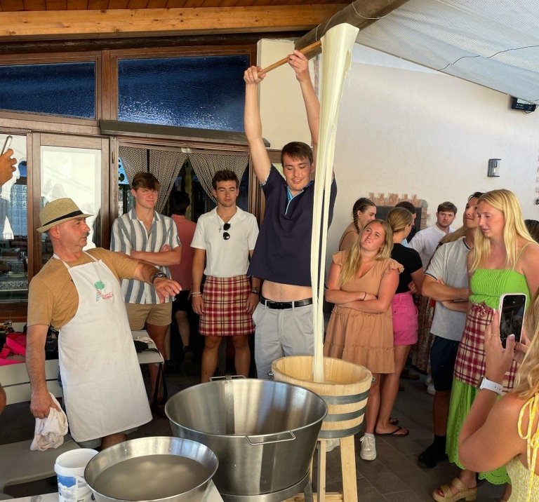 A man lifts a paddle high with mozzarella cheese stretched from a bucket. A man with an apron and surrounding group watch.
