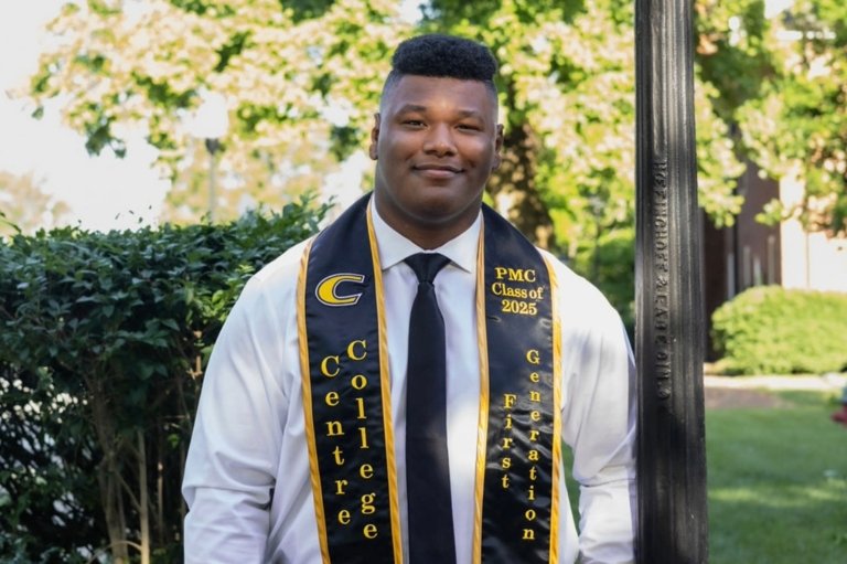 A headshot of a man leaning against a light pole. He wears a white shirt, tie and black graduation stole.