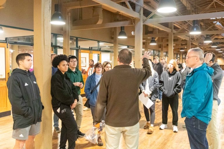 A large group of college students and adults stand in the middle of a large central room with laboratory spaces seen in the background.