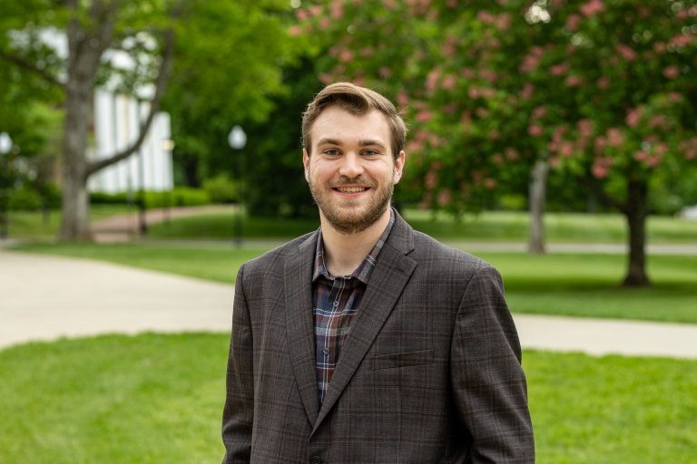 A young man in a dark blazer poses for a photo in front of a green lawn filled with trees. 