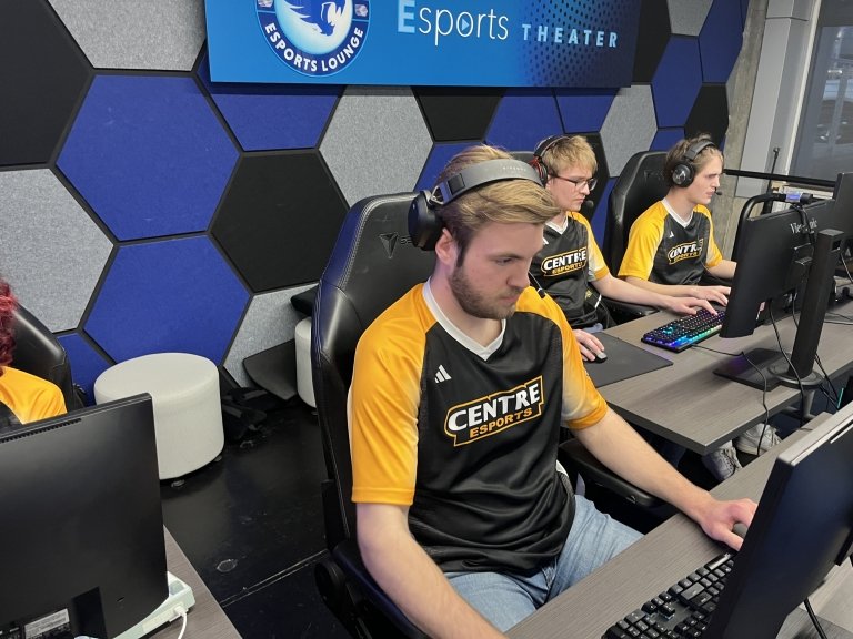 A young man in a black and gold Centre College Esports jersey sits at a computer competing in a tournament. 
