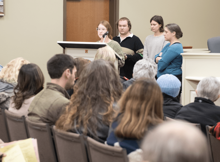 A young woman speaks into a microphone at a lectern in Danville City Hall as students and a professor stand behind her and a seated audience looks on. 