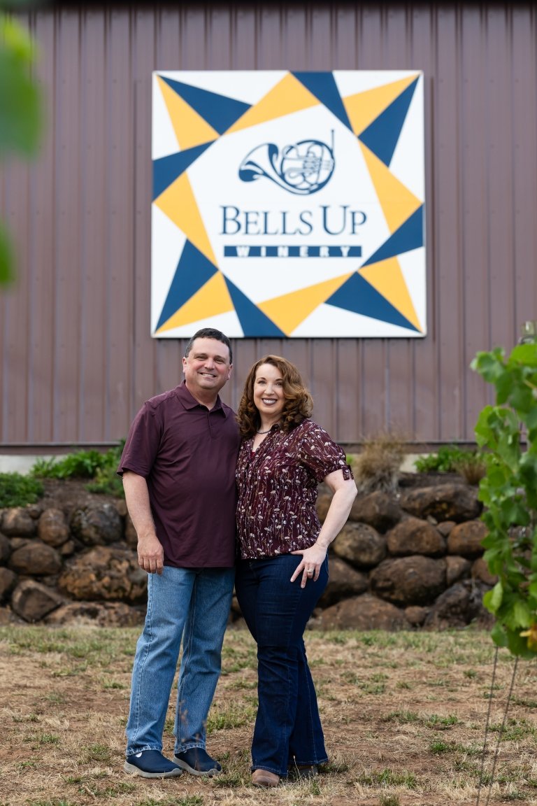 A man and woman stand side-by-side on front of a stone and metal building with a sign reading "Bells Up Winery" mounted to the side. 