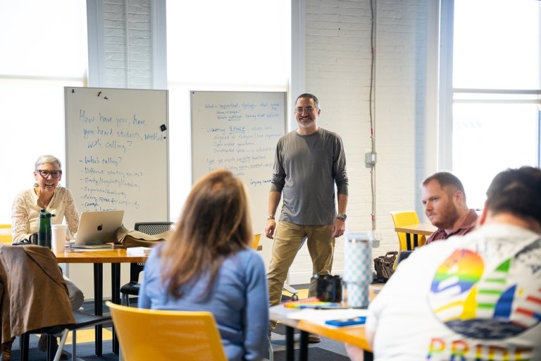 A man standing at the front of a room in front of two white boards filled with notes smiles as a woman seen in the foreground addresses the group. 