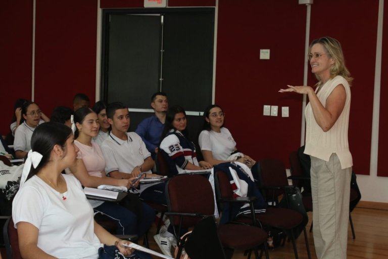 A woman stands in front of a room of people sitting at desks as she shares a lesson.