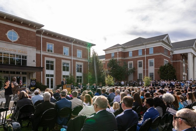 A large crowd sits in front of two large brick buildings as speakers offer remarks at a podium.