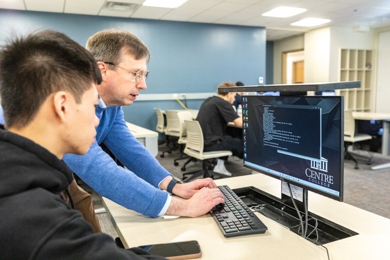 Centre College professor Thomas Allen, wearing a blue sweater, leans over a computer to help a student in his AI class. 