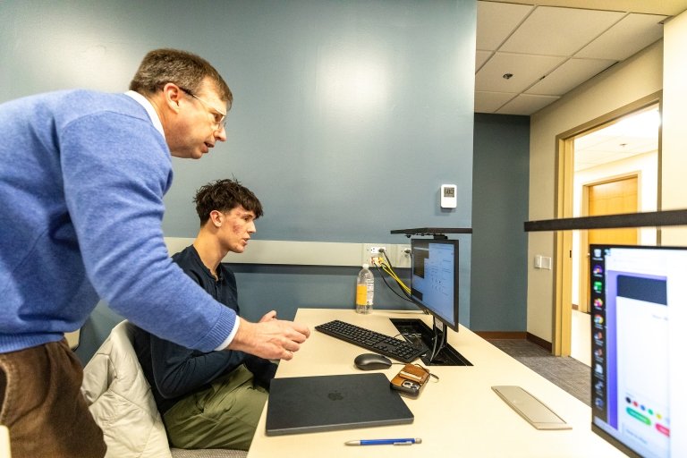 Centre College professor Thomas Allen, wearing a blue sweater, leans over a computer to help a student in his AI class. 