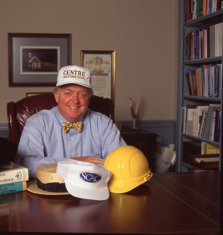 In a file photo, former Centre College President Michael F. Adams sits at his desk, wearing a Centre cap with a gold hard hat and a Centre visor on the desk in front of him. 