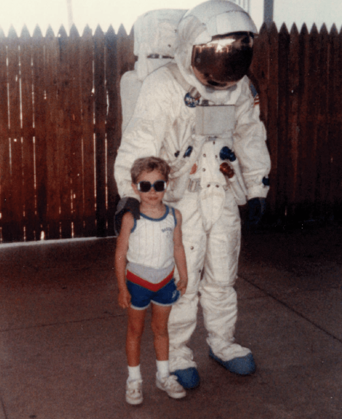 A young boy, Centre grad Jimmy Skaggs, wearing a a tank top, shorts and large sunglasses, poses next to someone in a 1980s era space suit&nbsp;