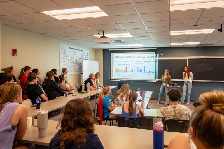 Two young woman stand at the front of a crowded classroom presenting their research to classmates and professors during the Centre College RICE Symposium.