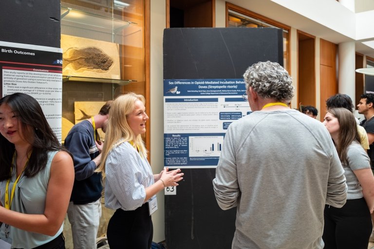 A young woman talks to a man while standing in front of a presentation poster in a hallway filled with other students during the Centre College RICE Symposium.