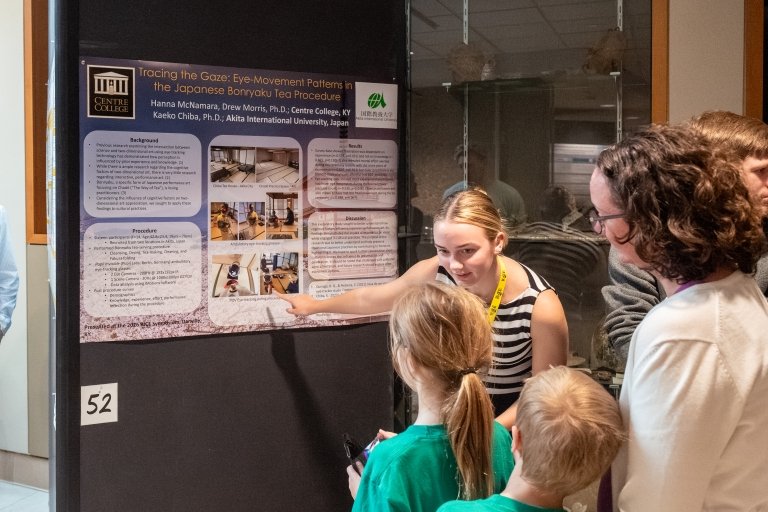 A young woman points to a research paper behind her as she leans forward to talk to two young girls standing with another woman enjoying the presentation.