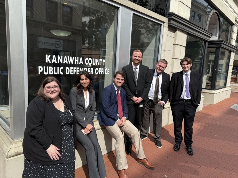 Four men and two women, all wearing business suits pose in front of a glass window which reads "Kanawa County Public Defender's Office."