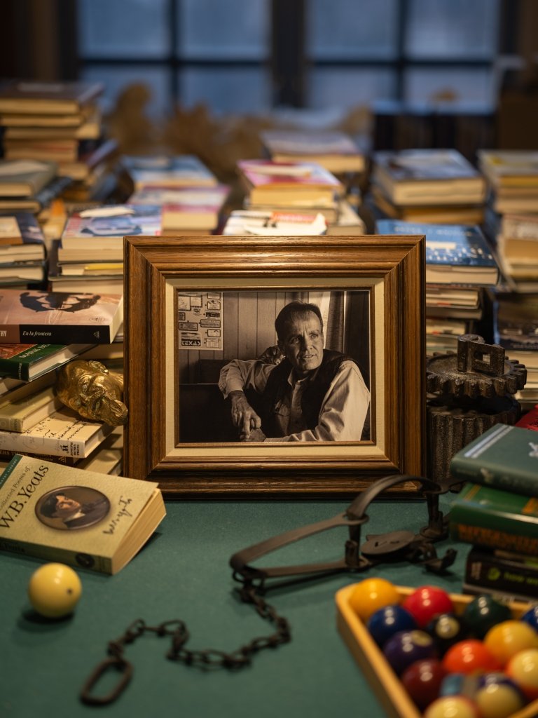 A framed back-and-white photo of American author Cormac McCarthy sits on a pool table covered with books.