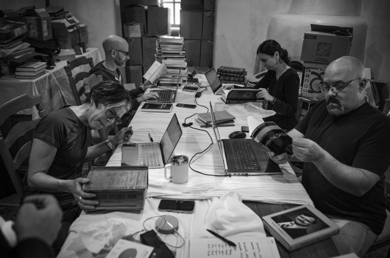 In a black-and-white photo, members of the Cormac McCarthy Society, including Centre College English professor Stacey Peebles sit around a rectangular table filled with books and papers in the home of author Cormac McCarthy.