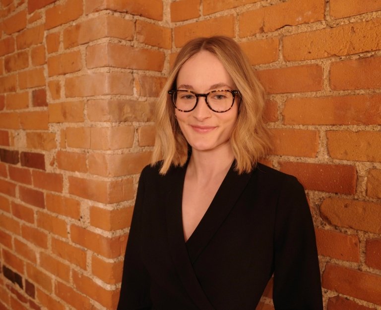 A young woman poses for a photo in a black business suit while standing in front of a brick wall.
