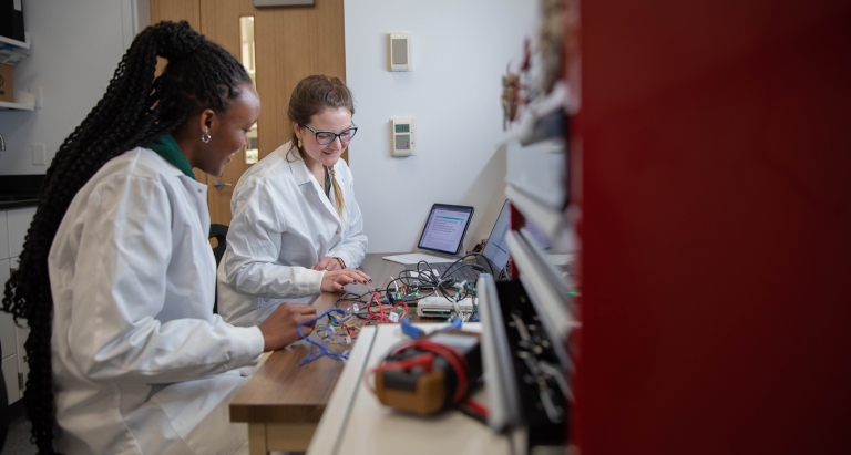 Two young women in white lab coats sit at a workstation covered with scientific devices.
