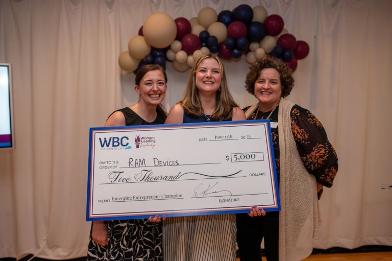 A young woman stands between two other women while holding a large ceremonial check for RAM Devices from the Women's Business Center of Kentucky in the amount of $5,000.