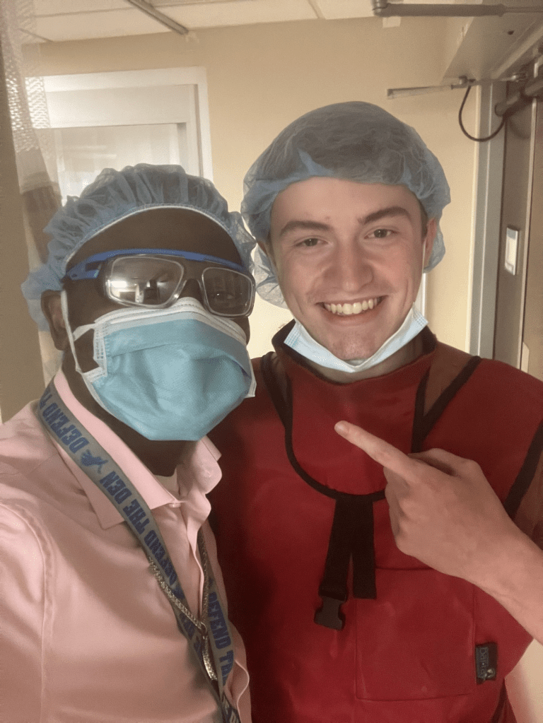 Christopher Ferguson smiles for a photo with the physician he's shadowing. They're both wearing hairnets and surgical masks.